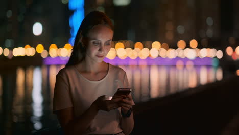At-night-a-young-girl-sits-on-the-grass-in-a-big-city-and-looks-into-the-smartphone-screen-writes-a-message-and-maintains-her-personal-blog.